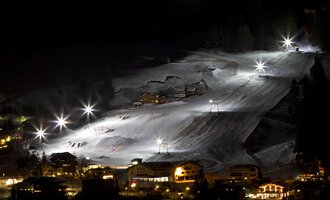 A lit ski slope at night with several skiers. In the foreground, huts and lights are visible. | © Sport Kessler GmbH