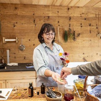 © Kleinwalsertal Tourismus - Bettina Kessler Eine lächelnde Frau in einer Holzküche reicht einer Person Blumen und kleine Flaschen. Im Hintergrund sind Kräuter und verschiedene Zutaten zu sehen. | © Kleinwalsertal Tourismus - Bettina Kessler