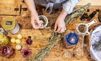 Eine Person bereitet Kräuter und blühende Pflanzen auf einem Holztisch vor. Neben ihnen stehen verschiedene Glasbehälter mit Zutaten und Utensilien für die Verarbeitung. | © Kleinwalsertal Tourismus - Bettina Kessler