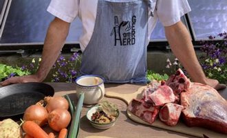 Ein Koch steht an einem Tisch mit frischen Zutaten, darunter Gemüse und Fleisch. Im Hintergrund sind Blumen und eine helle Umgebung sichtbar. | © Gerd am Herd | Gerd Hammerer