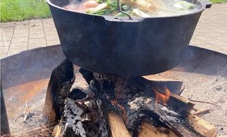 Ein Kessel mit frischen Zutaten über einem offenen Feuer. Im Hintergrund sind grüne Hügel und Bäume zu sehen. | © Gerd am Herd | Gerd Hammerer