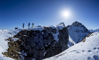 A group of hikers stands on a snowy summit. The sun shines brightly over the mountains. | © Kleinwalsertal Tourismus eGen | Dominik Berchtold