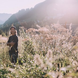 © Das Naturhotel Chesa Valisa****s Ein junger Mann steht in einem blühenden Feld mit hohen Gräsern. Die Sonne scheint und die Berge sind im Hintergrund sichtbar. | © Das Naturhotel Chesa Valisa****s
