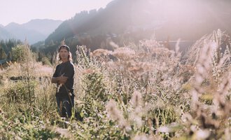 Ein junger Mann steht in einem blühenden Feld mit hohen Gräsern. Die Sonne scheint und die Berge sind im Hintergrund sichtbar. | © Das Naturhotel Chesa Valisa****s