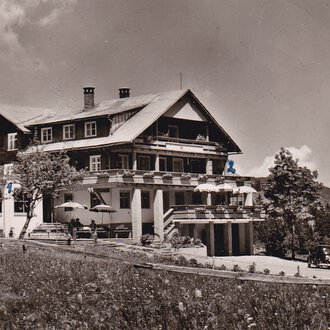 © Genuss- & Aktivhotel Sonnenburg Ein historisches Hotel in den Bergen, umgeben von Bäumen und Wiesen. Im Vordergrund sind alte Autos und eine Terrasse mit Sonnenschirmen zu sehen. | © Genuss- & Aktivhotel Sonnenburg