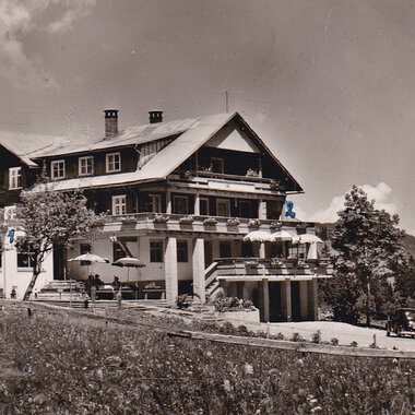Ein historisches Hotel in den Bergen, umgeben von Bäumen und Wiesen. Im Vordergrund sind alte Autos und eine Terrasse mit Sonnenschirmen zu sehen. | © Genuss- & Aktivhotel Sonnenburg