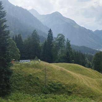© Nicole Lughammer Eine malerische Landschaft mit grünen Hügeln und hohen Bergen. Im Vordergrund steht eine kleine Hütte umgeben von Bäumen. | © Nicole Lughammer