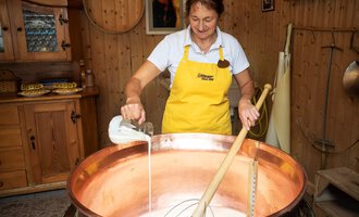 A woman in a yellow apron is stirring in a large copper pot with milk. In the background, wooden shelves and a cozy decor can be seen. | © Schönegger Käsealm
