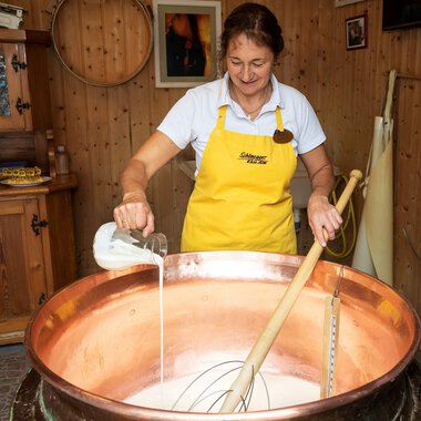 A woman in a yellow apron is stirring in a large copper pot with milk. In the background, wooden shelves and a cozy decor can be seen. | © Schönegger Käsealm