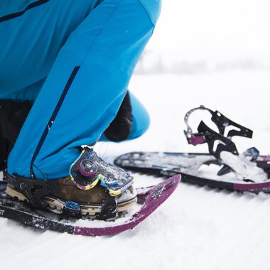 Eine Person schnallt Schneeschuhe an, um im Schnee zu wandern. Die Umgebung ist winterlich und schneebedeckt. | © Kleinwalsertal Tourismus | Oliver Farys