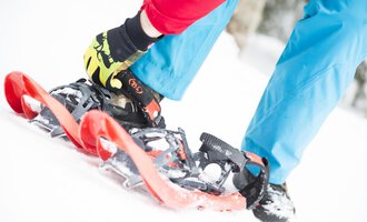 Eine Person schnallt sich Schneeschuhe an, während sie im Schnee steht. Der Boden ist weiß und die Kleidung ist bunt. | © Kleinwalsertal Tourismus eGen | Oliver Farys