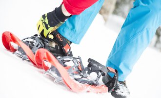 Eine Person schnallt Schneeschuhe an, während sie auf Schnee steht. Die Person trägt eine rote Jacke und blaue Hose, was einen farbenfrohen Kontrast zur winterlichen Umgebung bildet. | © Kleinwalsertal Tourismus eGen | Oliver Farys