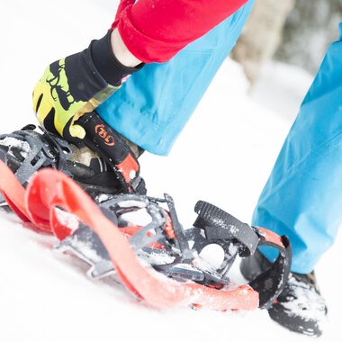 Eine Person schnallt Schneeschuhe an, während sie auf Schnee steht. Die Person trägt eine rote Jacke und blaue Hose, was einen farbenfrohen Kontrast zur winterlichen Umgebung bildet. | © Kleinwalsertal Tourismus eGen | Oliver Farys