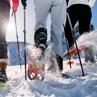 Three people are hiking in the snow with snowshoes. The sun is shining and snow is spraying from their movements. | © Kleinwalsertal Tourismus | Oliver Farys