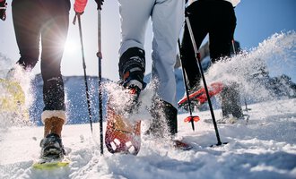 Three people are hiking in the snow with snowshoes. The sun is shining and snow is spraying from their movements. | © Kleinwalsertal Tourismus | Oliver Farys
