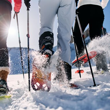 Three people are hiking in the snow with snowshoes. The sun is shining and snow is spraying from their movements. | © Kleinwalsertal Tourismus | Oliver Farys