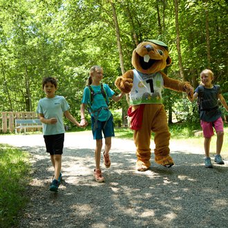 © Kleinwalsertal Tourismus | Oliver Farys Kinder gehen fröhlich auf einem Weg durch den Wald, begleitet von einem lustigen Maskottchen. Die Atmosphäre ist freundlich und einladend. | © Kleinwalsertal Tourismus | Oliver Farys