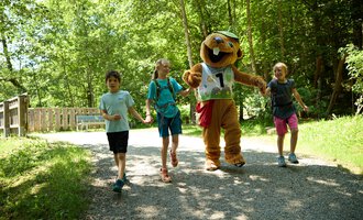 Children are happily walking along a path through the forest, accompanied by a cheerful mascot. The atmosphere is friendly and inviting. | © Kleinwalsertal Tourismus | Oliver Farys