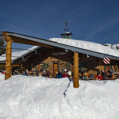 A cozy mountain cabin in the snow with guests on the terrace. The sky is clear and blue, and the surroundings are wintry. | © Bergstüble | Michaela Beck Dornach