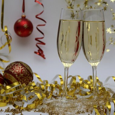 Zwei Sektgläser stoßen auf einem festlich dekorierten Tisch an. Im Hintergrund sind rote Weihnachtskugeln und goldene Dekoration zu sehen. | © Genuss- & Aktivhotel Sonnenburg
