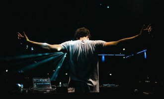 A DJ stands behind his mixing console with outstretched arms. Colorful lights shine in the background, creating a festive atmosphere. | © dj let´s fetz