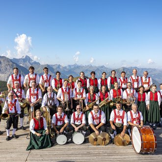 © Trachtenkapelle Riezlern Eine Musikkapelle in traditioneller Tracht steht auf einer Plattform in den Bergen. Im Hintergrund sind beeindruckende Berge und blauer Himmel zu sehen. | © Trachtenkapelle Riezlern