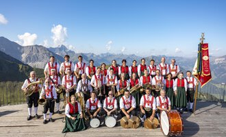Eine Musikkapelle in traditioneller Tracht steht auf einer Plattform in den Bergen. Im Hintergrund sind beeindruckende Berge und blauer Himmel zu sehen. | © Trachtenkapelle Riezlern
