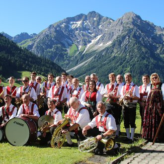 Eine Musikkapelle in traditioneller Tracht steht vor einer malerischen Berglandschaft. Im Hintergrund sind grüne Wiesen und schneebedeckte Berge zu sehen. | © Musikverein d´Hirschegger | Jockl Metzger