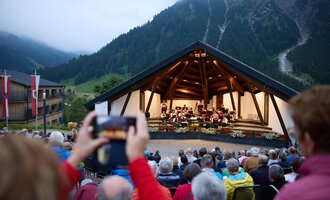 Eine Freiluftbühne in den Bergen mit einer Orchesteraufführung. Viele Zuschauer sitzen davor und genießen die Musik in der Abenddämmerung. | © Kleinwalsertal Tourismus | Oliver Farys
