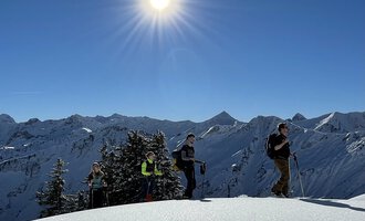 A group of hikers stands on a snow-covered summit under bright sunlight. In the background, snow-covered mountains can be seen. | © Abenteuer Vertical | Christian Kohler
