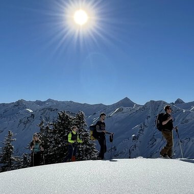 A group of hikers stands on a snow-covered summit under bright sunlight. In the background, snow-covered mountains can be seen. | © Abenteuer Vertical | Christian Kohler