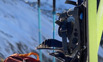 A sled and a ski bag are lying in the snow in front of a mountainous landscape. In the background, ski poles are visible. | © Abenteuer Vertical | Christian Kohler