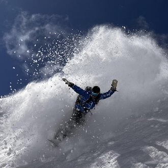 A snowboarder rides through fresh snow, creating a spray cloud. The sky is clear and blue, ideal for winter sports. | © Abenteuer Vertical | Christian Kohler