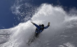 A snowboarder rides through fresh snow, creating a spray cloud. The sky is clear and blue, ideal for winter sports. | © Abenteuer Vertical | Christian Kohler