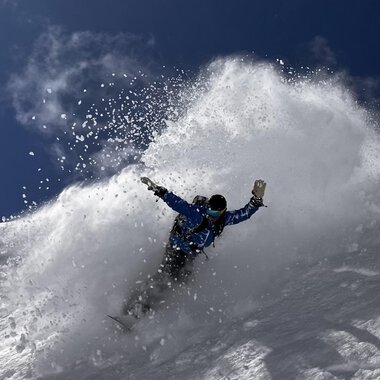 A snowboarder rides through fresh snow, creating a spray cloud. The sky is clear and blue, ideal for winter sports. | © Abenteuer Vertical | Christian Kohler