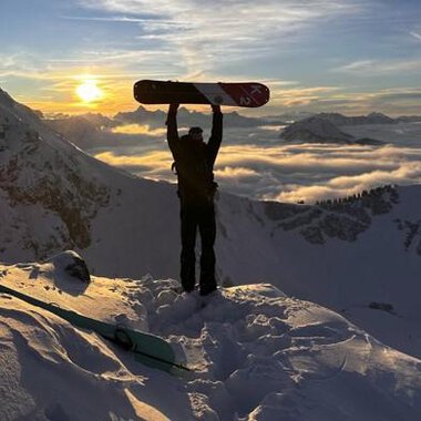 A snow sports enthusiast stands on a snowy peak, holding a snowboard up high. In the background, the sun shines between the mountains and a blanket of clouds glows in golden colors. | © Abenteuer Vertical | Christian Kohler