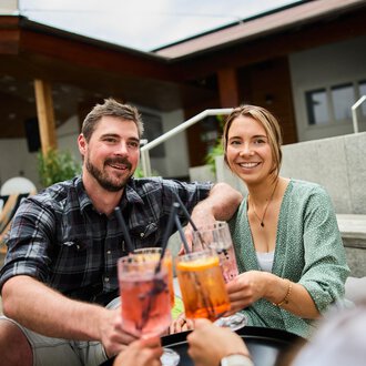 © Kleinwalsertal Tourismus | Oliver Farys Ein fröhliches Paar, das zusammen Cocktails genießt. Im Hintergrund sind gemütliche Sitzgelegenheiten sichtbar. | © Kleinwalsertal Tourismus | Oliver Farys