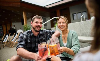 Ein fröhliches Paar, das zusammen Cocktails genießt. Im Hintergrund sind gemütliche Sitzgelegenheiten sichtbar. | © Kleinwalsertal Tourismus | Oliver Farys