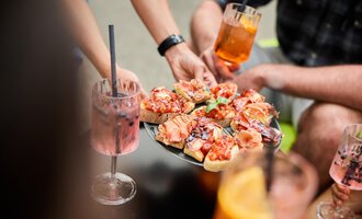 EinTablett mit Antipasti, garniert mit Schinken und Tomaten. Umgeben von Getränken und Menschen, die eine gesellige Zeit genießen. | © Kleinwalsertal Tourismus | Oliver Farys