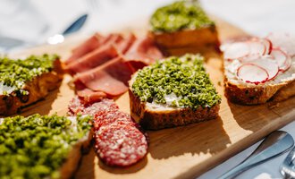 Ein hübsches Holzbrett mit verschiedenen belegten Broten. Die Snacks sind mit grünen Kräutern, Schinken und Radieschen dekoriert. | © Kleinwalsertal Tourismus eGen | Oliver Farys
