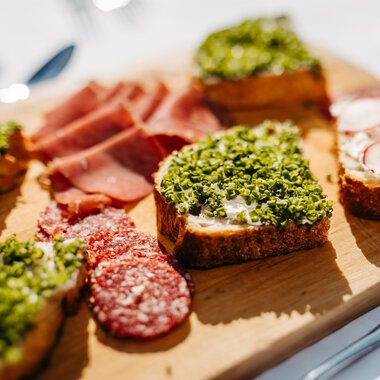 Ein hübsches Holzbrett mit verschiedenen belegten Broten. Die Snacks sind mit grünen Kräutern, Schinken und Radieschen dekoriert. | © Kleinwalsertal Tourismus eGen | Oliver Farys