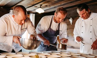 Drei Köche arbeiten konzentriert in einer rustikalen Küche. Sie bereiten Gerichte in weißen Schalen vor und verwenden verschiedene Kochutensilien. | © Kleinwalsertal Tourismus eGen | Oliver Farys