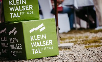 Grüne Würfeln mit dem Schriftzug "Klein Walsertal seit 1302". Im Hintergrund sind Stühle und Tische in einem Außenbereich sichtbar. | © Kleinwalsertal Tourismus eGen | Oliver Farys