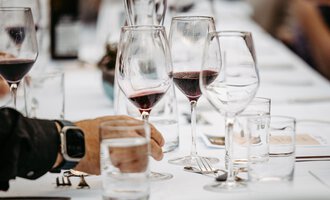 Ein elegant gedeckter Tisch mit Weingläsern und Wasserkrügen. Im Vordergrund ist eine Hand zu sehen, die ein Wasserglas hält. | © Kleinwalsertal Tourismus eGen | Oliver Farys