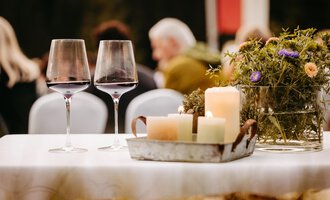 Ein schön gedeckter Tisch mit zwei Weingläsern und einer rustikalen Dekoration. Im Hintergrund sind Gäste auf einer Veranstaltung zu sehen. | © Kleinwalsertal Tourismus eGen | Oliver Farys