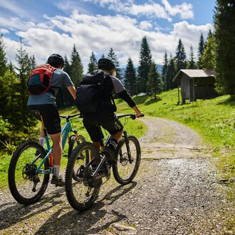 © Kleinwalsertal Tourismus | Oliver Farys Zwei Radfahrer fahren einen Weg entlang in einer grünen Landschaft. Im Hintergrund sind Bäume und eine kleine Hütte zu sehen. | © Kleinwalsertal Tourismus | Oliver Farys