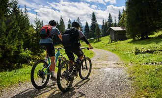 Zwei Radfahrer fahren einen Weg entlang in einer grünen Landschaft. Im Hintergrund sind Bäume und eine kleine Hütte zu sehen. | © Kleinwalsertal Tourismus | Oliver Farys
