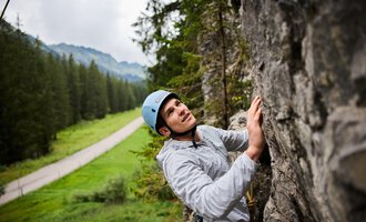 Ein Kletterer, der an einer Felswand arbeitet, trägt einen Helm und konzentriert sich auf seinen Aufstieg. Im Hintergrund sind Bäume und eine Straße in einer grünen Landschaft zu sehen. | © Bergschule Kleinwalsertal | Oliver Farys