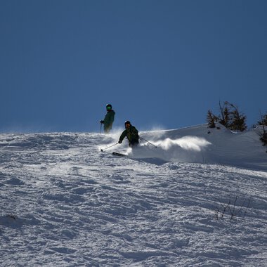 Zwei Skifahrer fahren über einen schneebedeckten Hang. Der Himmel ist klar und blau. | © Markus Hoefler