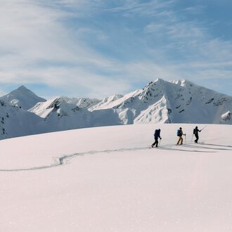 A skier in a blue suit is skiing through fresh, powdery snow. In the background, snow-covered mountains and a clear blue sky can be seen. | © Kleinwalsertal Tourismus | Oliver Farys